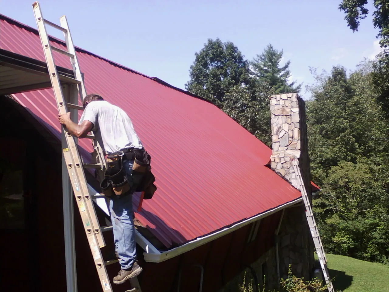 Roofer on a ladder beside a red metal roof and stone chimney — Soffit & Fascia Repair by Roofing Brothers in Rutherfordton